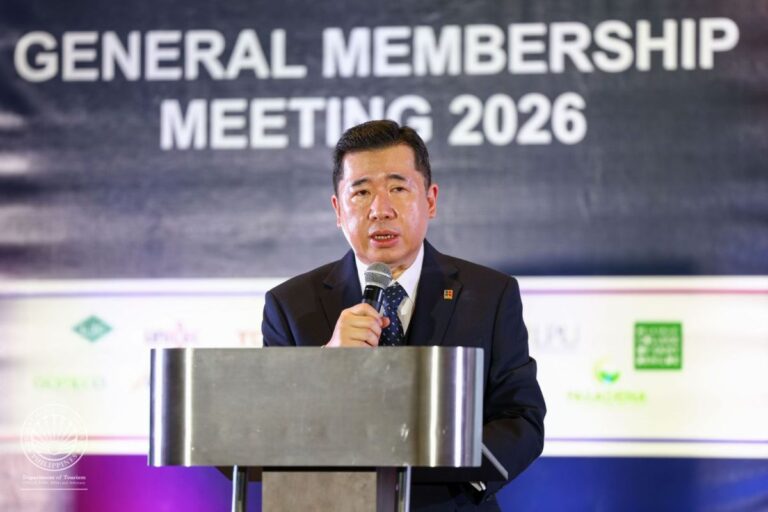 Asian man in a suit speaks into a microphone at a podium on stage during the General Membership Meeting 2026.
