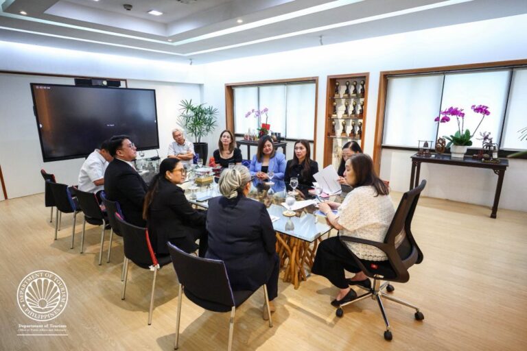 A diverse group of professionals seated around a glass conference table in a modern meeting room, documents and glasses on the table, a large screen on the wall, and a Philippines Department of Tourism logo in the corner.