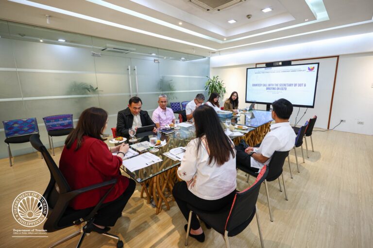 Group of professionals seated around a glass conference table, looking at a presentation on a large screen in a bright, modern meeting room.