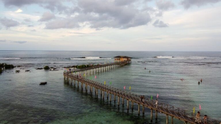 Long wooden pier extends over turquoise water toward a thatched hut at the end, with people swimming nearby and colorful flags along the railing.