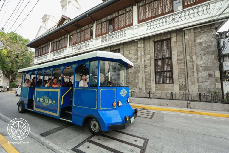 Blue open-sided tourist tram filled with passengers, traveling along a street beside a historic stone building marked Intramuros.