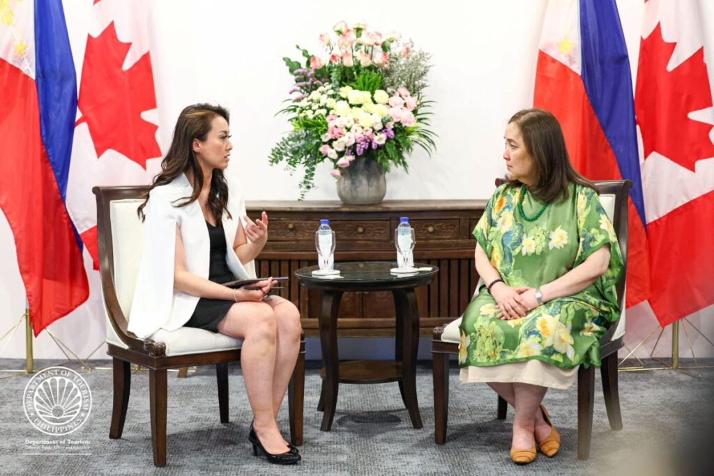 Two women in a formal interview setting, seated in armchairs with a round table and water bottles between them; flags and a large flower arrangement visible behind.