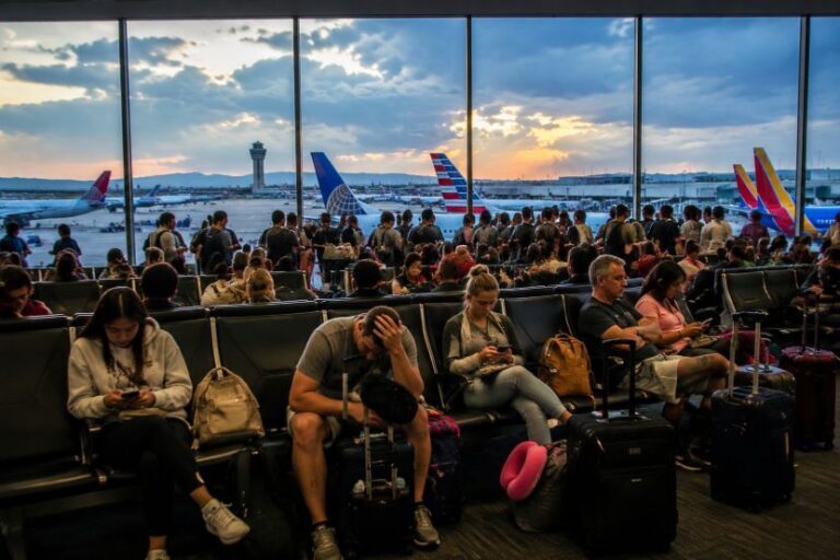 Passengers seated in an airport terminal with luggage, many looking at phones as planes wait on the runway outside at sunset.