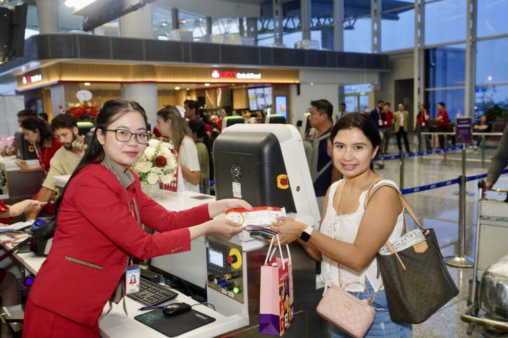Airport check-in counter: smiling staff in red uniform hands a boarding pass to a passenger at a self-service kiosk.