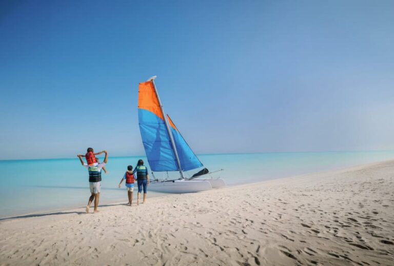 Family wearing life jackets approach a colorful sailboat on a white-sand tropical beach with turquoise water.