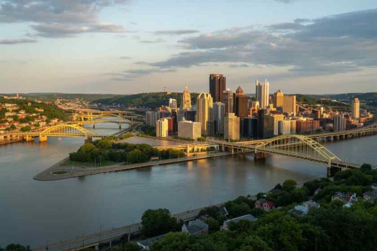 Aerial view of downtown Pittsburgh at sunset with yellow bridges spanning the river and surrounding hills.