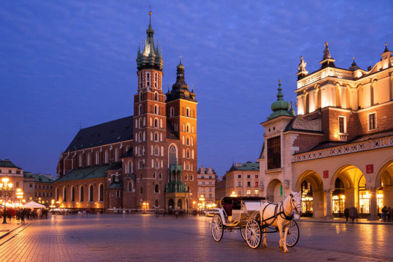 Evening view of Kraków's main square with St. Mary's Basilica and a white horse-drawn carriage in the foreground, warm streetlights glowing.