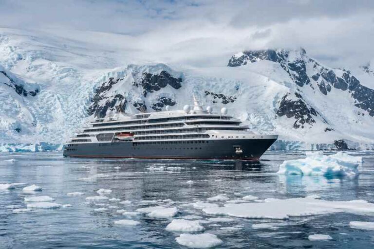 Large luxury cruise ship sailing through icy Arctic waters with snow-covered mountains in the background and ice floes nearby.
