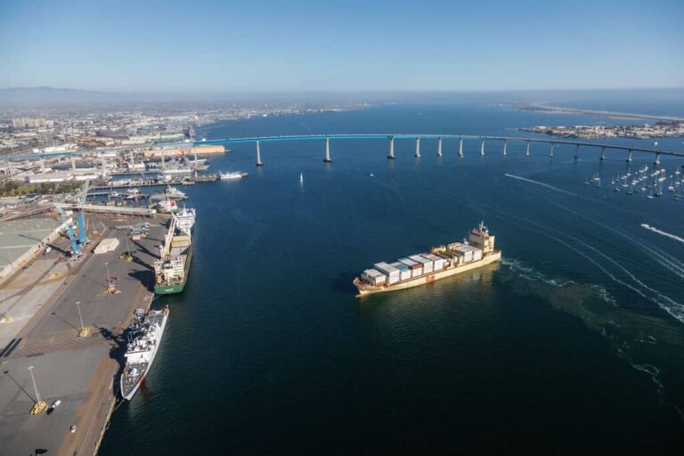 Aerial view of a harbor with a container ship, cargo cranes, and a long blue bridge over calm water.