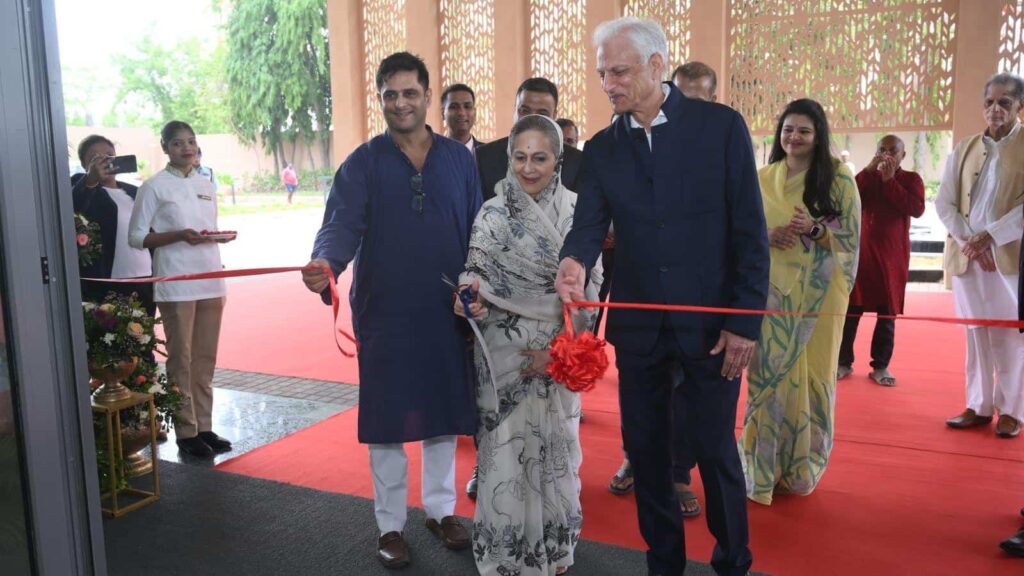 A ribbon-cutting ceremony with three people at the front: a man in a blue kurta, a woman in a patterned saree, and a tall man in a dark suit, cutting a red ribbon.