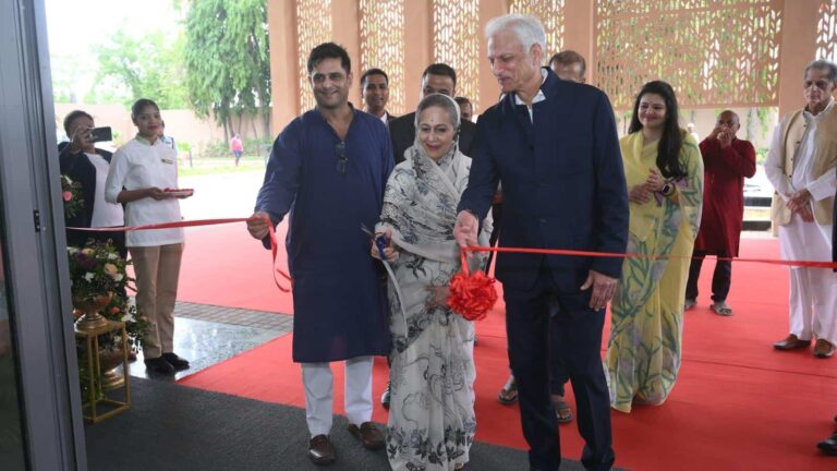 A ribbon-cutting ceremony with three people at the front: a man in a blue kurta, a woman in a patterned saree, and a tall man in a dark suit, cutting a red ribbon.
