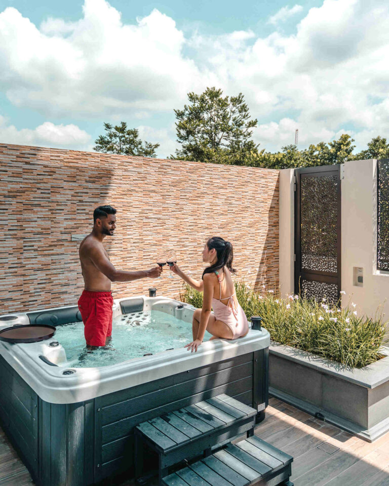 Couple toasting with wine glasses in a backyard hot tub beside a brick privacy wall and plants.