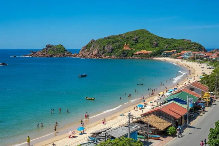 Sunlit tropical beach with clear blue water, sandy shore, and green hills in the background; people swim and stroll near colorful beachside buildings.
