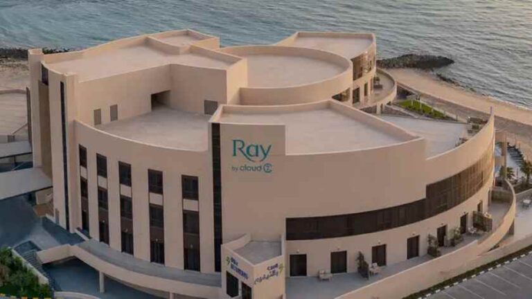 A large curved beige hotel building by the beach, with a sign that reads 'Ray by Cloud' on the facade, seen from an aerial angle.