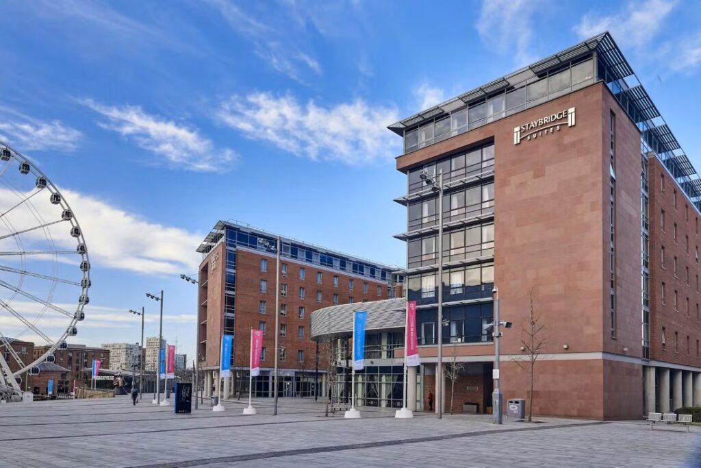 Modern Staybridge Suites hotel in a brick building, set in a spacious plaza with a large Ferris wheel on the left and blue sky above.