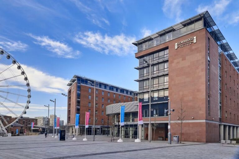 Modern Staybridge Suites hotel in a brick building, set in a spacious plaza with a large Ferris wheel on the left and blue sky above.
