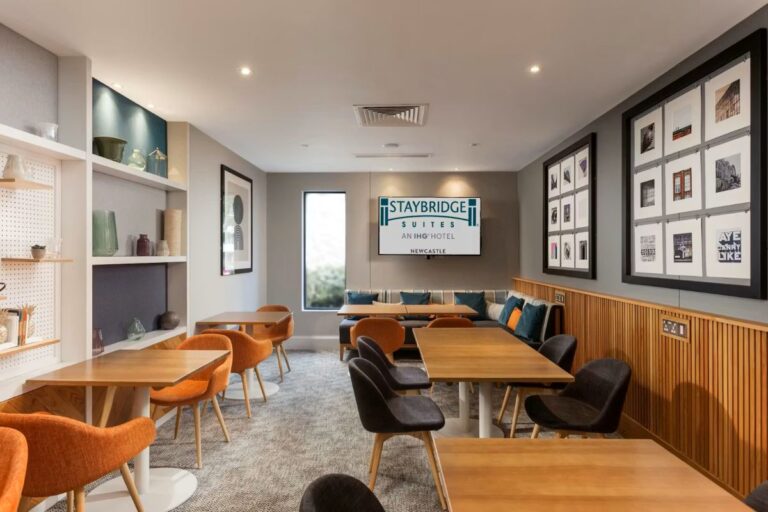 Hotel lounge at Staybridge Suites Newcastle with wooden tables, orange and black chairs, a gray sectional with teal cushions, wall art, and a Staybridge sign on a screen at the back.