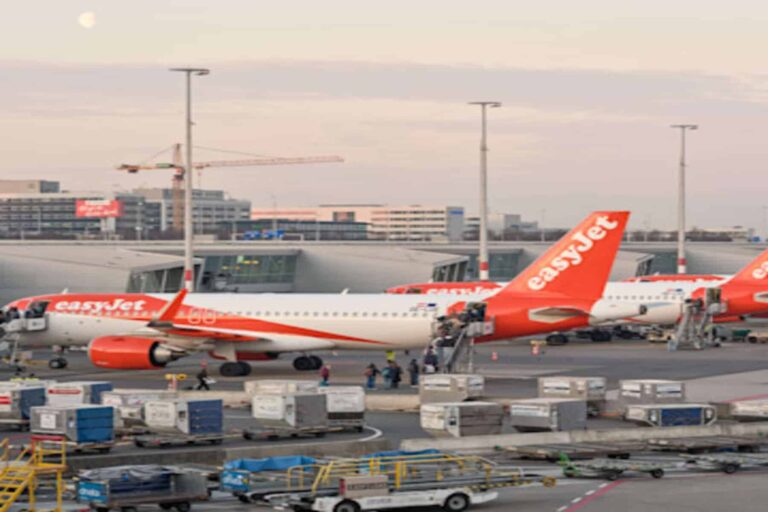 Several orange EasyJet airplanes parked at a terminal on the tarmac with ground crews and luggage carts nearby at dusk/early evening window edge.