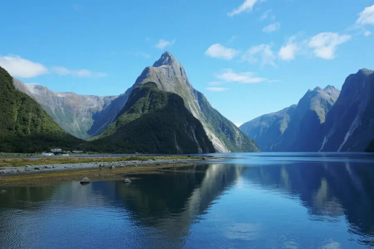 Jagged mountain rising over a calm blue fjord, forested slopes, and a rocky shoreline reflected in the water.