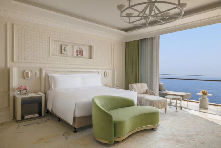 Luxurious hotel bedroom with a white bed, pale walls, and a green curved lounge chair, opening to a sea-view balcony.