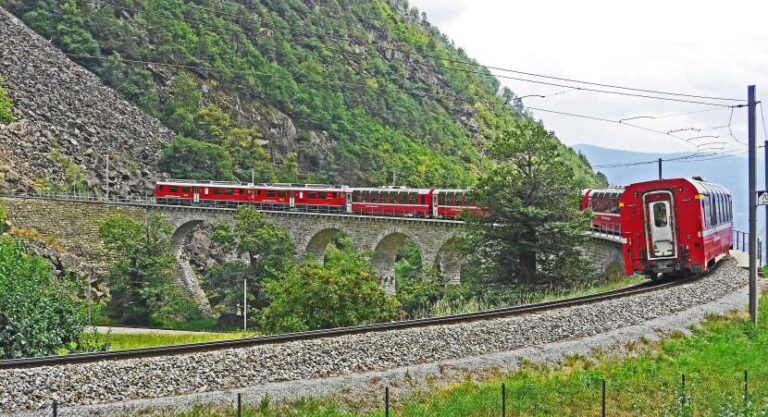 Red passenger train winding along a stone arched viaduct in a green mountainside landscape.