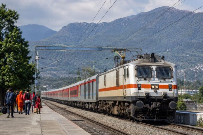 A passenger train at a platform with people waiting, set against green mountains and blue sky in the background.