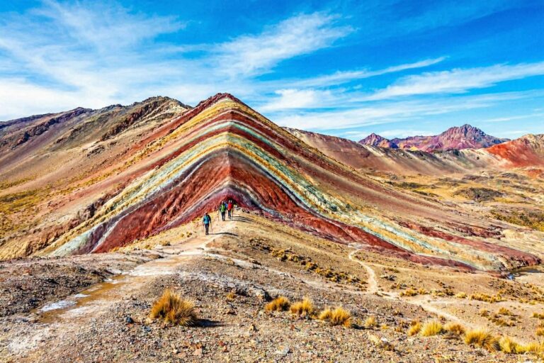 Colorful layered ridge mountain with hikers walking along a dirt trail in a desert landscape under a blue sky.