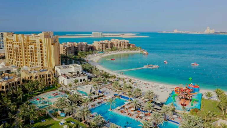 Aerial view of a tropical resort with a sandy beach, turquoise water, and palm-lined pool areas.