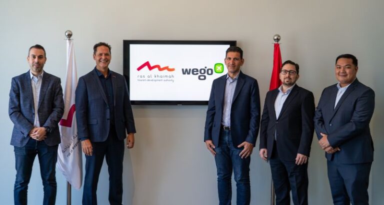Five business professionals stand side by side in front of a wall with a screen displaying logos and two flags nearby.