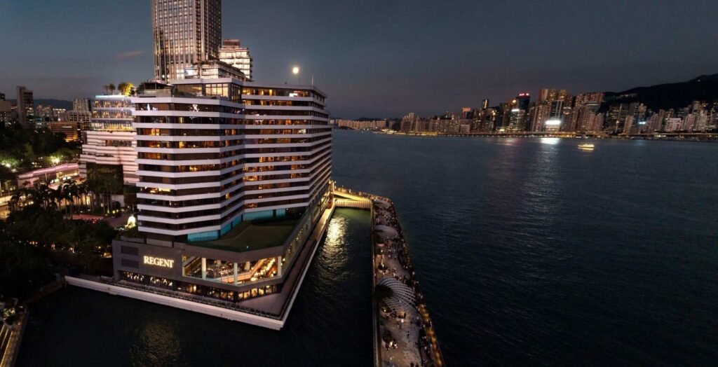 Nighttime aerial view of a waterfront city with a curved, illuminated Regent hotel building and a lit promenade along the water horizon across the bay.