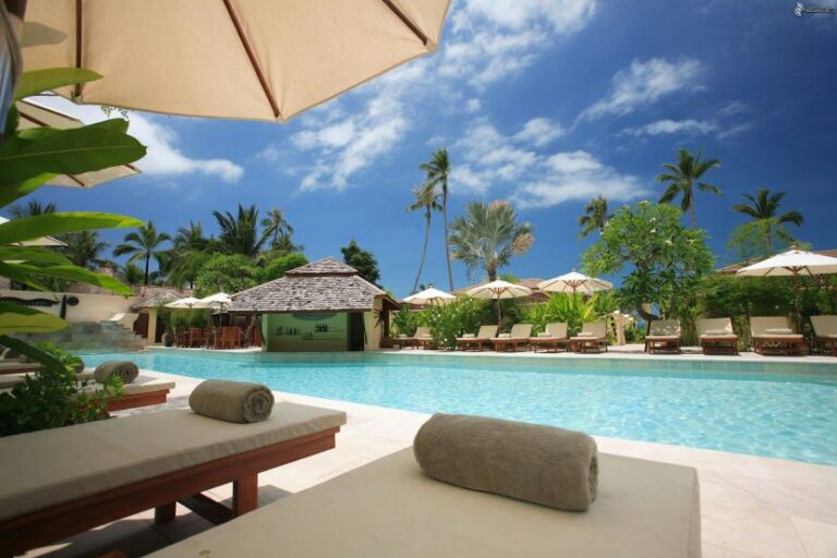 Resort pool area with cushioned lounge chairs, beige umbrellas, and a thatched poolside bar under a blue sky.