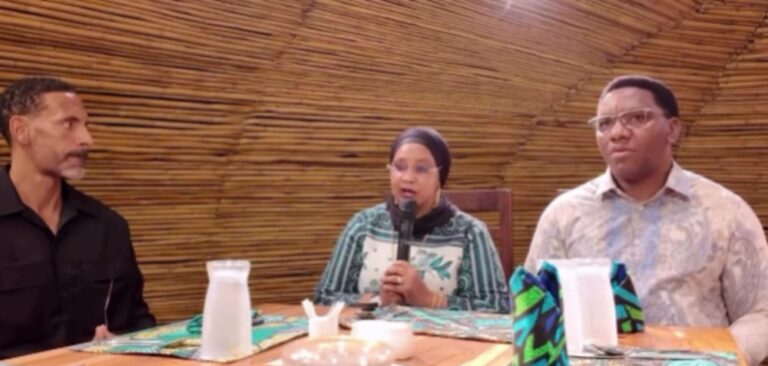 Three people sit at a wooden table; a woman in the center speaks into a microphone while two men listen nearby.