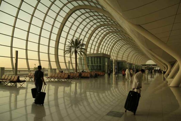 Interior of a modern airport terminal with curved glass arches, palm trees, and travelers pulling suitcases.