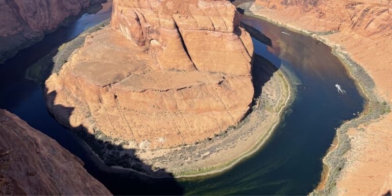 Aerial view of a red rock canyon with a horseshoe-shaped river bend; a small white boat is on the water near the shore.