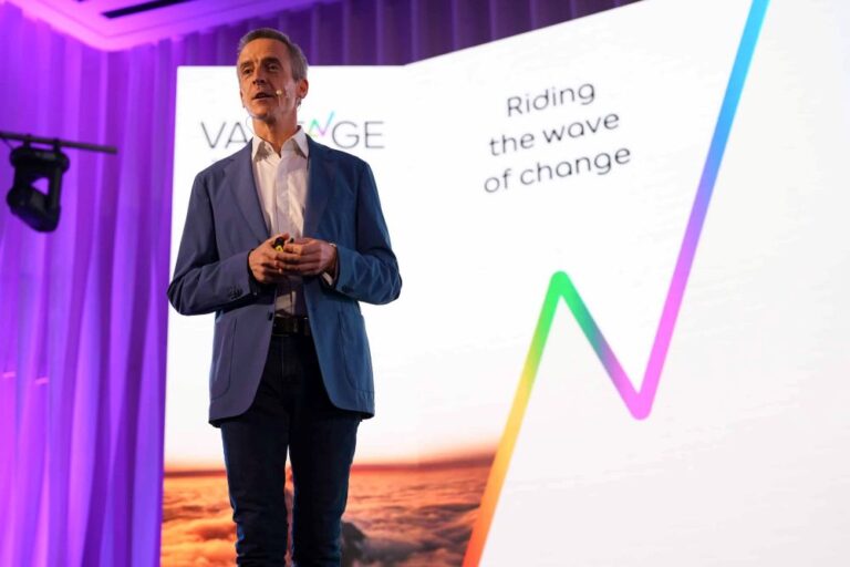 Male speaker in a blue blazer presenting on stage with a large screen showing the slogan 'Riding the wave of change'.] ,