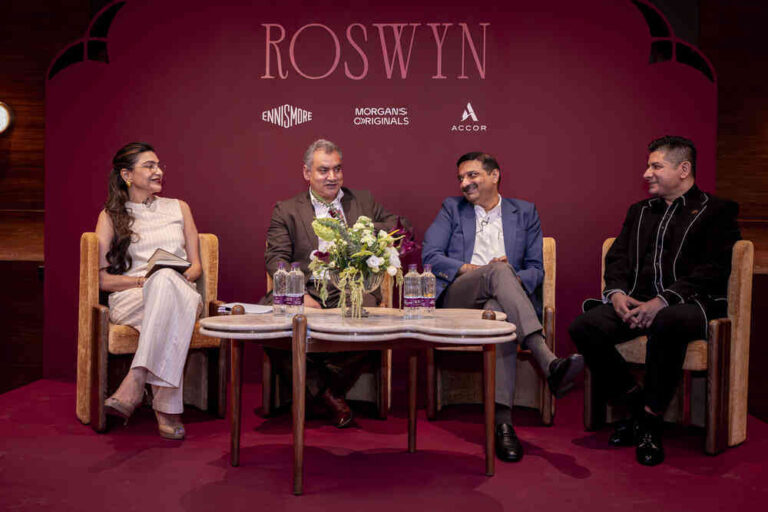 Panel discussion with four people seated on stage, maroon backdrop, and a flower centerpiece on the table below them.
