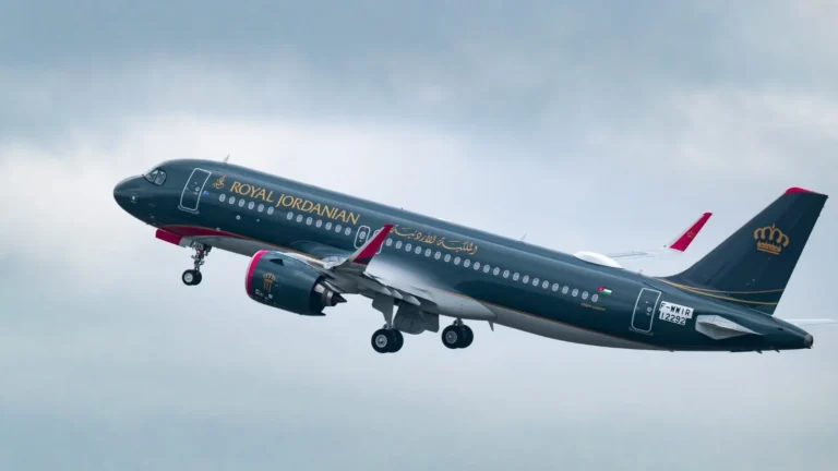 Royal Jordanian passenger aircraft in dark blue livery with gold lettering taking off against a cloudy sky, engine with pink accents visible under the wing, crown emblem on the tail.