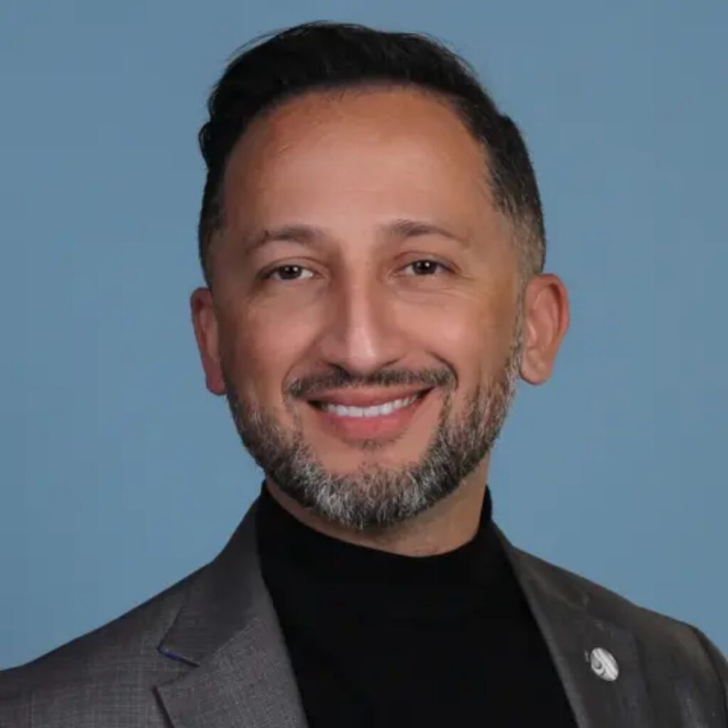 Head-and-shoulders portrait of a smiling man with short dark hair and a salt-and-pepper beard, wearing a gray blazer over a black turtleneck, against a blue background.