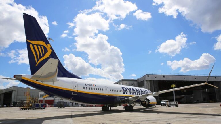 Ryanair plane on the tarmac with blue-and-yellow livery in front of a hangar, under a cloudy blue sky