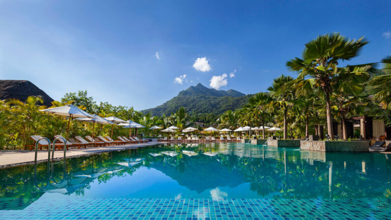 Resort swimming pool lined with lounge chairs and umbrellas, tropical trees, and a mountain range under a clear blue sky in the background.