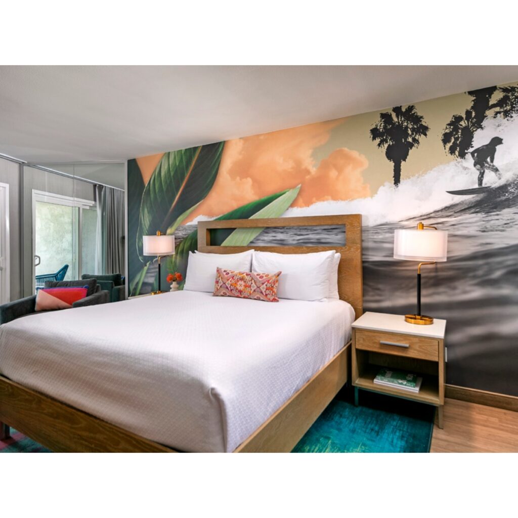 Bedroom with a large wooden bed, white bedding, and tropical mural featuring waves and palm silhouettes behind; two matching nightstands and lamps flank the bed.