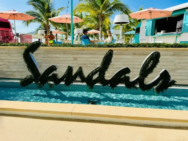 Large cursive sign reading 'sandals' covered in greenery sits in a shallow pool at a tropical resort, with palm trees and pink umbrellas behind it.