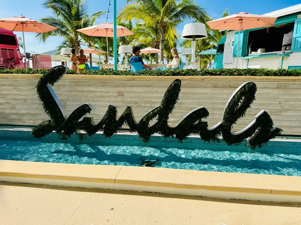 Large cursive hedge sign spelling 'sandals' over a shallow pool at a tropical resort, with palm trees, umbrellas, and a tiki-style counter in the background.