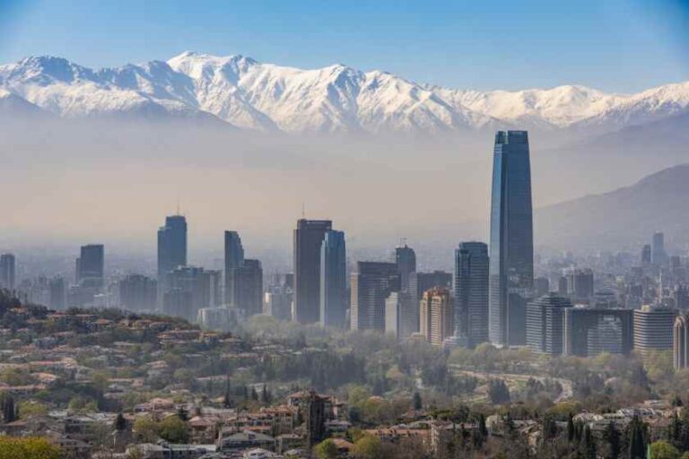 City skyline with tall skyscrapers and snow-capped mountains in the distance, under a clear blue sky.