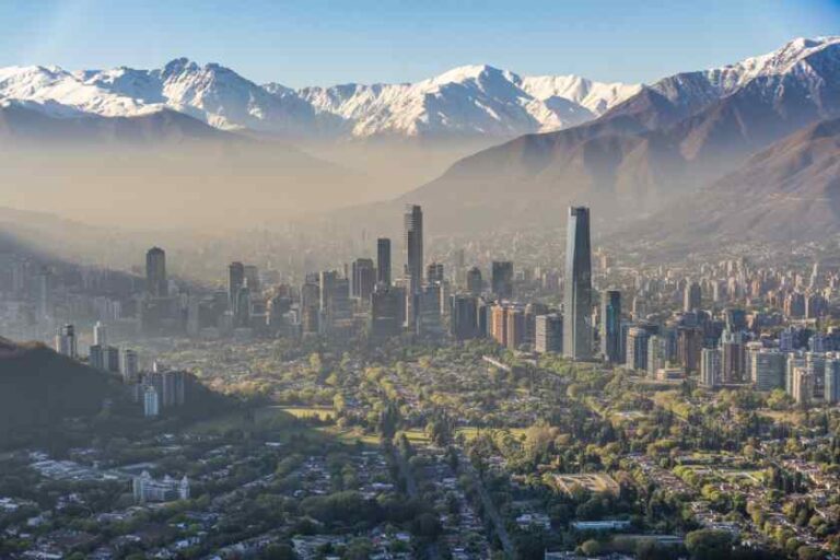 Aerial view of a city skyline with tall skyscrapers and green suburbs, mountains with snow in the distance and a hazy atmosphere