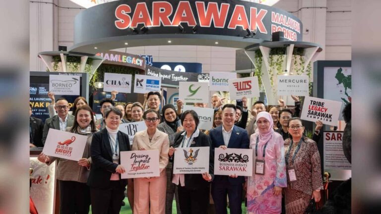 Group of diverse businesspeople at a travel expo holding signs for Sarawak and partner brands, smiling at the camera.
