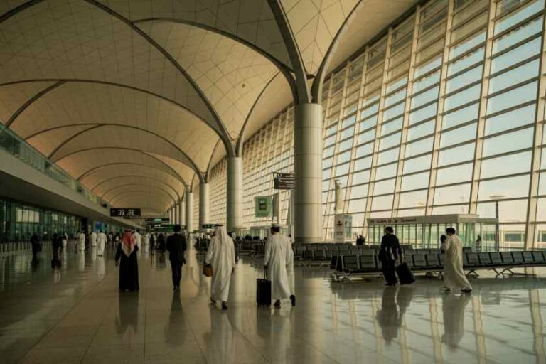 Spacious airport terminal concourse with travelers walking and luggage under a high curved ceiling and large glass walls.