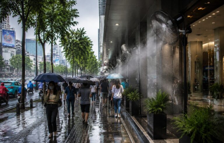 Pedestrians with umbrellas walk along a wet city sidewalk beside a glass-fronted building on a rainy day.