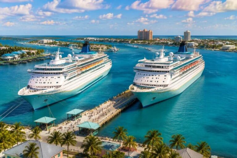 Two large white cruise ships docked at a sunny tropical port with palm trees and turquoise water.