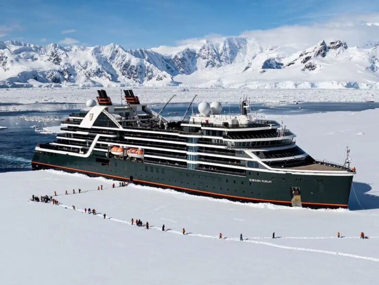 Polar cruise ship on a snow-covered sea with passengers walking ashore against rugged icy mountains and blue sky.